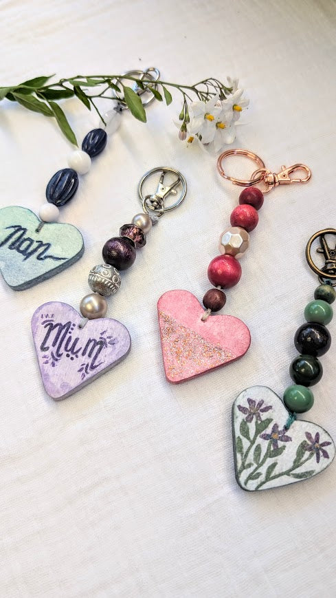 A collection of four handmade keyrings, each featuring a hand-painted heart pendant and a beaded string. The keyrings display soft pastel tones with unique designs: one says “Nan” in blue, another says “Mum” in purple, one is pink with gold shimmer, and the last features delicate purple flowers. They are laid out on a white cloth beside a sprig of chamomile flowers.
