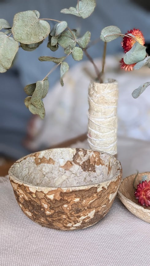 A handcrafted large paper bowl with a textured, earthy brown and cream design is displayed on a neutral cloth surface. Behind it sits a small vase wrapped in natural twine, holding dried eucalyptus leaves and red strawflowers. A smaller paper dish with dried flowers is partially visible to the right, creating a warm, natural aesthetic.
