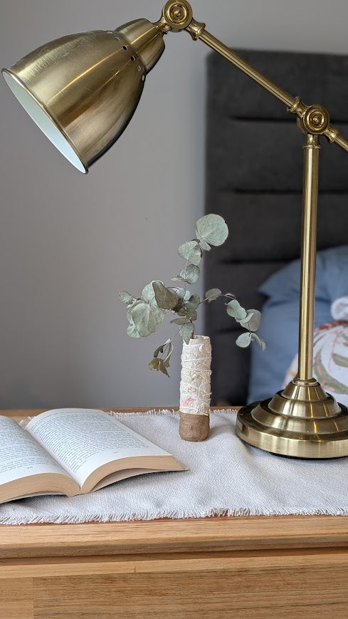 cozy bedside table scene featuring a brass adjustable desk lamp, an open book, and a small handmade paper clay vase holding dried eucalyptus stems. The vase has a natural brown base and a textured cream upper half with faint floral detailing. The table is covered with a neutral woven cloth, and a neatly made bed with soft pillows is visible in the background.