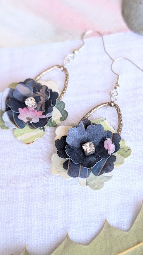 Handmade layered paper flower earrings with dark and pastel petals, sparkling centre stones, and gold teardrop hoops, displayed on a white fabric background.