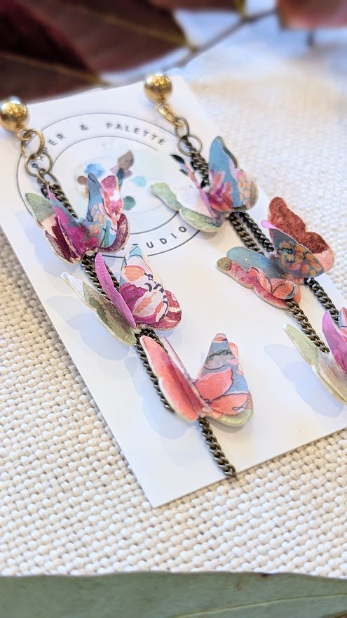 Close-up angled view of handcrafted butterfly earrings showing layered paper wings and brass chain details. Displayed on a product card against a textured fabric backdrop.