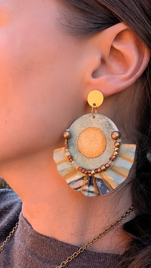 “Woman wearing statement paper earrings with fan-shaped pleats, warm sunlight highlighting hand-painted aqua and gold design.”