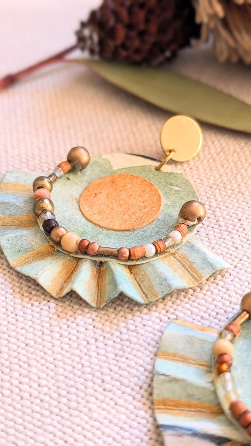 Close-up of single hand-painted paper earring with golden centre, brass stud, and warm-toned beading, styled with eucalyptus leaf