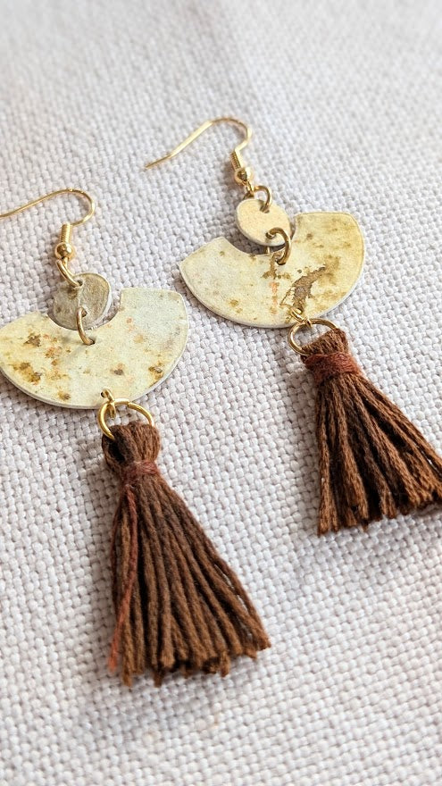 Close-up of handmade tassel earrings with brown cotton fringe, fan-shaped paper components, and gold hooks on textured fabric background