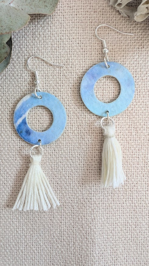 Flat lay of handmade round hoop earrings painted in soft blue and violet watercolour tones, with cream cotton tassels, displayed on natural linen fabric alongside eucalyptus leaves and coral.