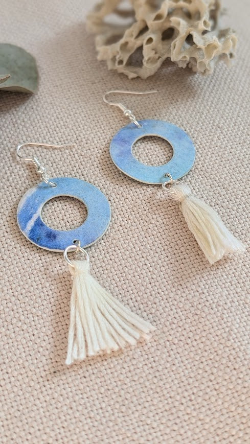 Flat lay of handmade round hoop earrings painted in soft blue and violet watercolour tones, with cream cotton tassels, displayed on natural linen fabric alongside eucalyptus leaves and coral.