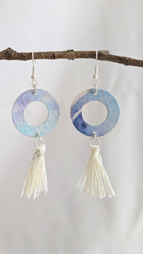 Close-up of blue and violet watercolour hoop earrings with cream cotton tassels, hanging from a natural twig against a soft white background.