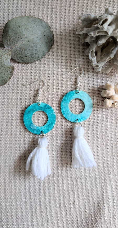 Flat lay of handmade aqua watercolour hoop earrings with white cotton tassels, styled on natural linen fabric with eucalyptus leaves and coral for a coastal, earthy look