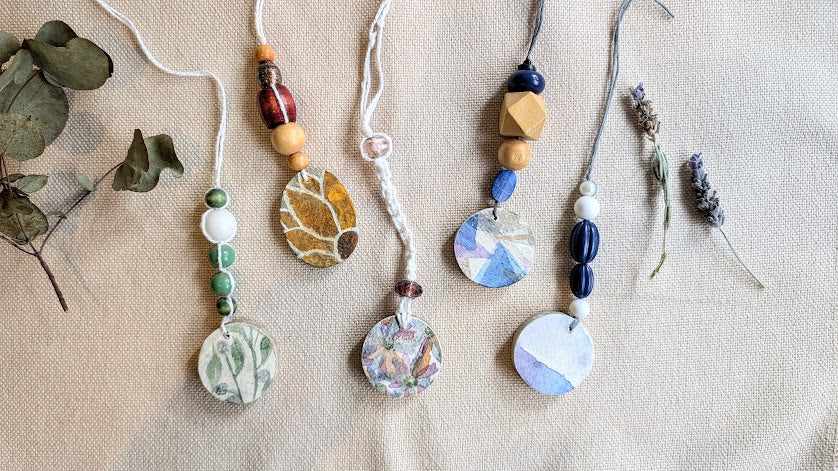 Handmade paper clay pendants on strings with natural elements on a textured fabric background