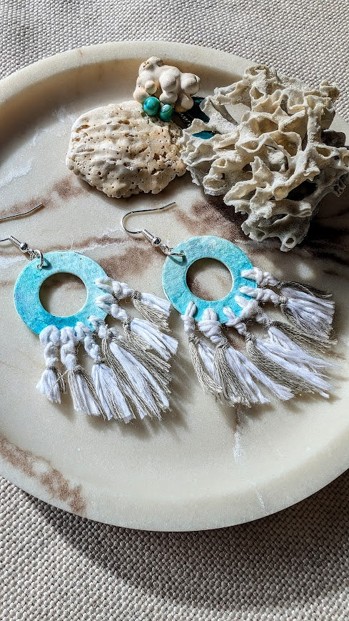 Pair of earrings with turquoise circular design and white tassels on a stone surface.