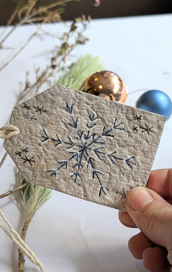 Handmade gift tag with snowflake design held by a hand, with decorative elements in the background.