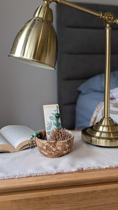 A cozy bedside scene featuring a brass desk lamp, an open book, and a decorative handmade paper bowl on a wooden nightstand. The bowl contains small natural items including a pine cone and green accessories, and a botanical-themed bookmark is propped up behind it. A soft fabric runner covers the top of the nightstand.