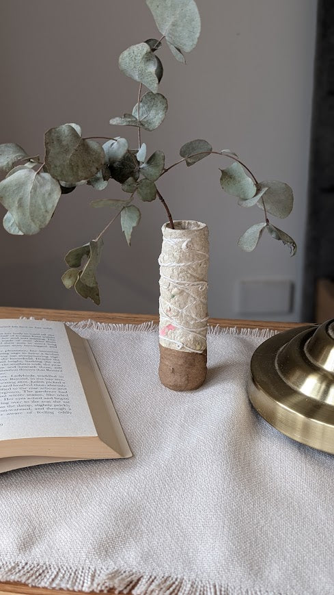 A small handmade paper clay vase with a natural brown base and cream textured upper half, featuring delicate floral details and wrapped string. It holds a sprig of dried eucalyptus and sits on a wooden bedside table next to an open book and a brass lamp, atop a neutral woven cloth.