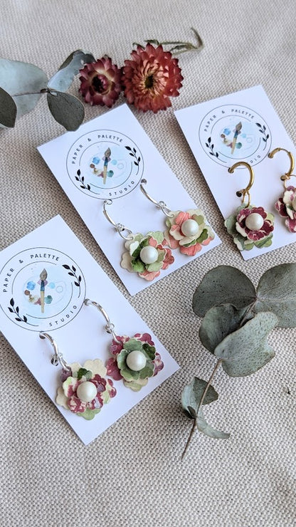 Flat lay of three pairs of handcrafted floral earrings from the Elowen Collection by Paper & Palette Studio. Each pair features layered pastel watercolour paper petals with a pearl centre, presented on branded earring cards. Surrounded by dried botanicals and eucalyptus leaves on a natural fabric background.