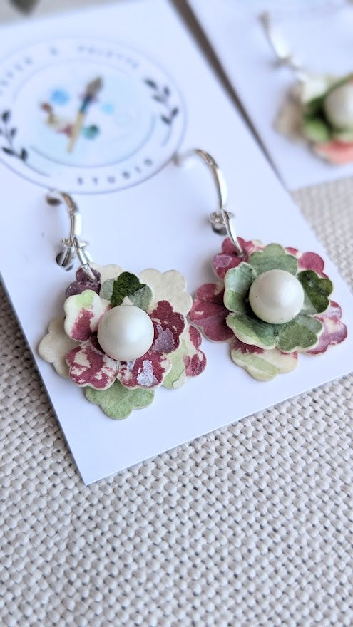 Close-up of a pair of handcrafted floral earrings from the Elowen Collection by Paper & Palette Studio. The earrings feature layered watercolour paper petals in soft green, blush, and burgundy tones with a central faux pearl, hanging from silver huggie hoops. Displayed on a textured cream fabric with a dried leaf for styling.