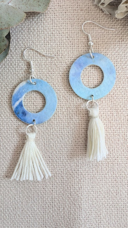 Flat lay of handmade round hoop earrings painted in soft blue and violet watercolour tones, with cream cotton tassels, displayed on natural linen fabric alongside eucalyptus leaves and coral.