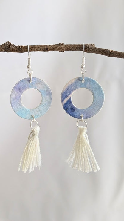 Close-up of blue and violet watercolour hoop earrings with cream cotton tassels, hanging from a natural twig against a soft white background.