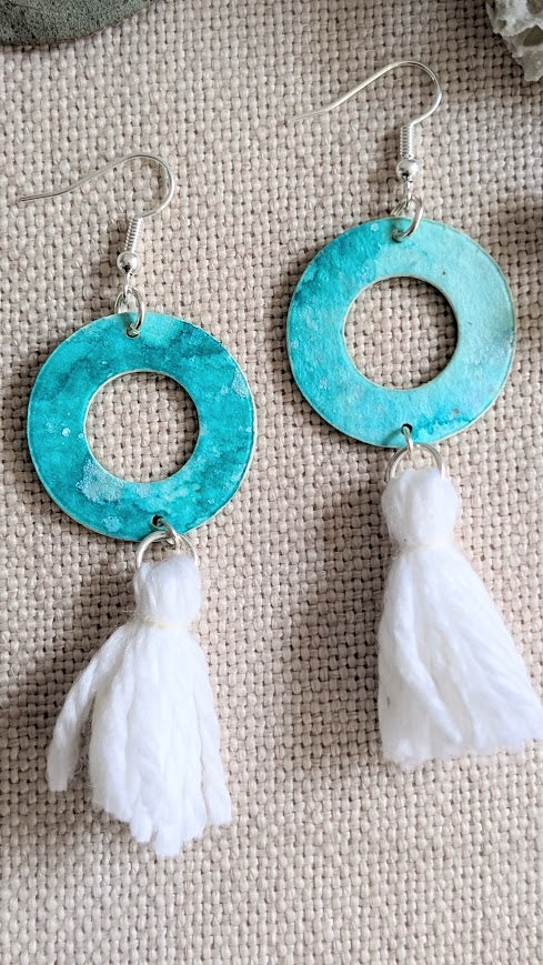 Close-up of artisan aqua hoop earrings with white tassels, hand painted in layered watercolour on a soft woven background.