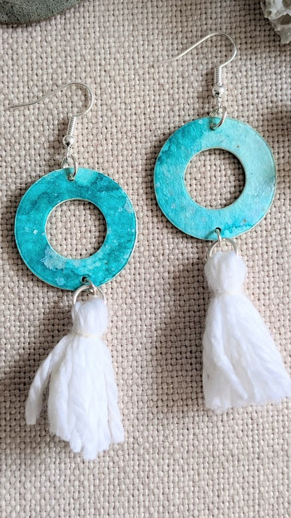 Close-up of artisan aqua hoop earrings with white tassels, hand painted in layered watercolour on a soft woven background.