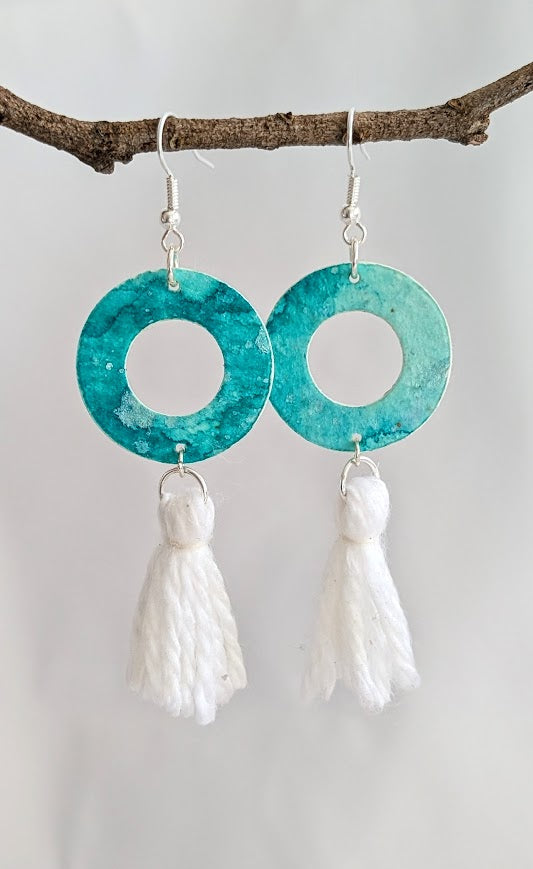 Pair of aqua watercolour hoop earrings with white cotton tassels, hanging from a natural twig against a clean white backdrop, showcasing their lightweight and artistic design.