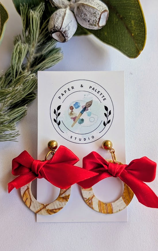 Earrings with red bows on a card from Paper & Palette Studio, surrounded by greenery.