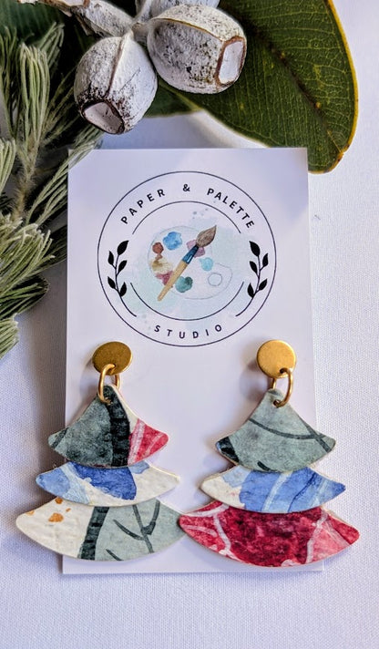 Colorful Christmas Tree earrings on a 'Paper & Palette Studio' branded card with green leaves in the background.