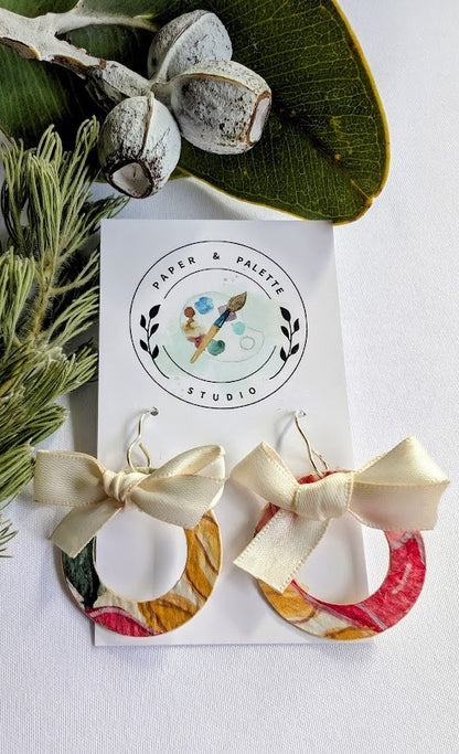 Colorful earrings with bows on a card from Paper & Palette Studio, surrounded by greenery.