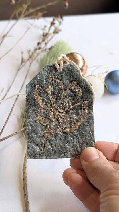 Hand holding a paper tag with a flower design on a white surface with dried plants and a Christmas decorations.