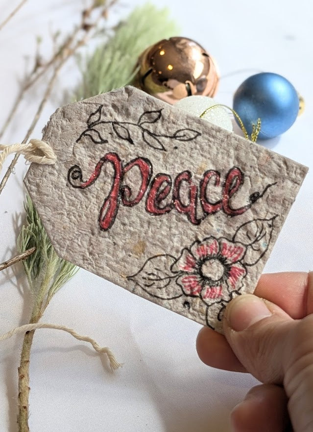 Handmade 'Peace' tag with floral designs held by a hand, surrounded by Christmas ornaments.