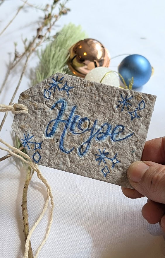 Handwritten 'Hope' tag with decorative elements, held over Christmas ornaments and branches.