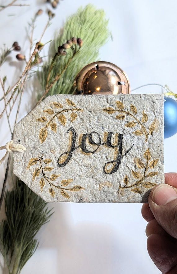 Decorative gift tag with 'joy' text and gold leaves, held against a blurred background with greenery and ornaments.